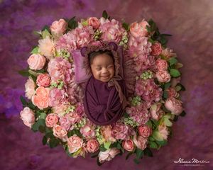 Smiling newborn baby wrapped in purple swaddle sleeping in heart-shaped arrangement of pink and peach roses.