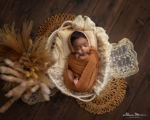Overhead view of newborn baby in orange swaddle sleeping in white bowl with lace and dried pampas grass.