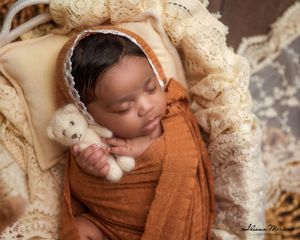 Newborn baby in rust orange swaddle and lace bonnet sleeping while holding a tiny teddy bear.