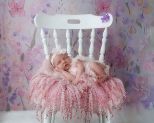 Peaceful newborn baby girl in pink lace dress sleeping on white chair with colorful floral wallpaper background.