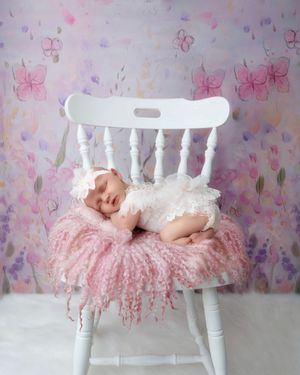 Sleeping newborn baby girl in white lace outfit on pink fluffy rug on white chair with floral backdrop.