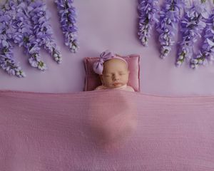 Newborn baby girl swaddled in pink wrap sleeping in miniature bed surrounded by purple wisteria flowers.