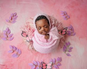 Newborn baby in pink wrap sleeping in round nest with butterflies and purple leaves on pink backdrop.