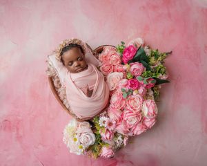 Newborn swaddled in pink wrap resting in heart-shaped bowl surrounded by pink and white roses.