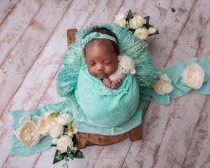 Newborn baby swaddled in teal wrap sleeping in wooden bowl with white flowers and floral headband.