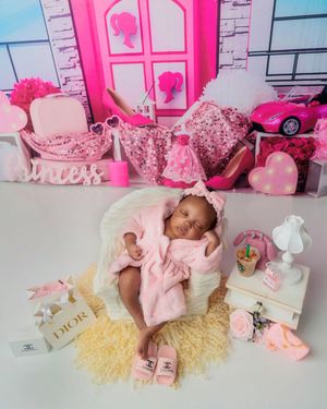 Sleeping newborn in pink robe posed in Barbie-themed setup with Dior and Chanel accessories.