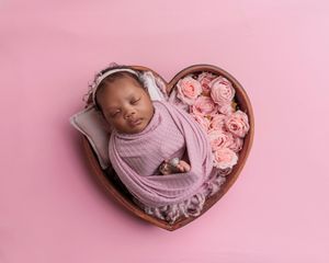 Sleeping newborn wrapped in pink swaddle inside heart-shaped wooden bowl with pink roses on pink background.