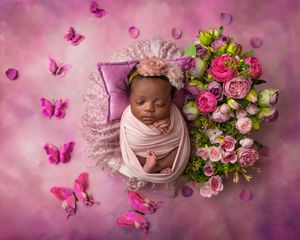 Newborn baby swaddled in pink wrap sleeping beside pink roses with butterflies on purple background.