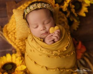 Newborn baby swaddled in mustard yellow wrap holding heart prop surrounded by sunflowers.