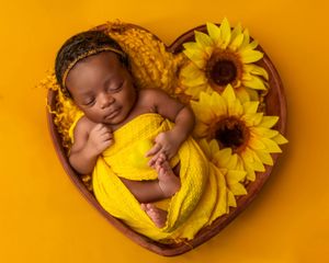 Newborn baby sleeping in heart-shaped bowl with sunflowers and yellow wrap on yellow background.
