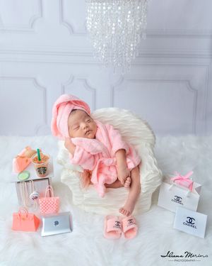 Newborn baby girl in pink robe sleeping surrounded by luxury brand miniature accessories.