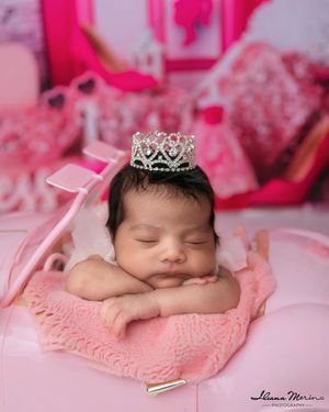 Sleeping newborn baby girl wearing crystal tiara posed in pink Barbie-themed newborn photography setup.
