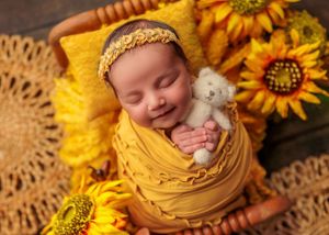 Smiling newborn baby wrapped in yellow swaddle holding teddy bear surrounded by sunflowers.