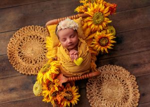 Sleeping newborn swaddled in yellow wrap surrounded by sunflowers on woven rattan mats.