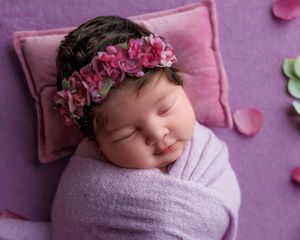 Peaceful newborn baby swaddled in lavender wrap with pink floral crown sleeping on purple pillow.