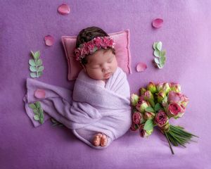 Sleeping newborn baby girl wrapped in lavender swaddled with floral headband on pink blanket with roses.