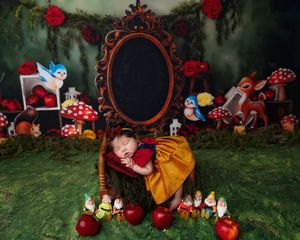 Sleeping newborn in Snow White costume posed in enchanted forest setting with dwarfs and red apples.