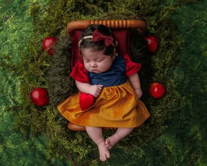 Newborn baby girl dressed as Snow White sleeping among red apples on lush green moss backdrop.