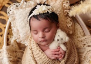 Sleeping newborn baby swaddled in cream knit wrap holding tiny teddy bear with lace bonnet.