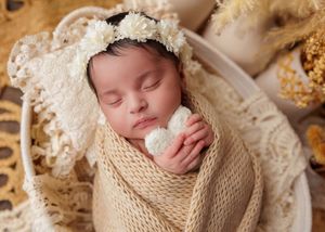 Newborn baby in cream knit swaddle and lace bonnet holding white flower in wicker basket.