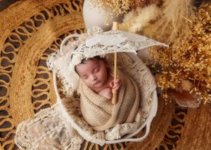 Sleeping newborn baby swaddled in beige wrap holding lace umbrella in white bowl, boho style.