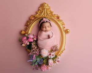 Sleeping newborn baby girl wrapped in pink swaddle inside an ornate gold frame with flowers.