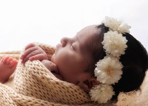 Sleeping newborn baby with white floral headband resting on cream knit blanket, side profile.