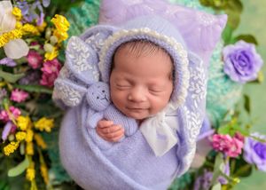 Smiling newborn in lavender wrap and bunny bonnet holding matching stuffed bunny.