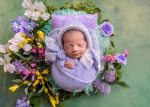 Sleeping newborn in lavender knit wrap and bonnet holding a stuffed bunny surrounded by colorful flowers.
