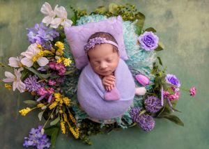 Peaceful newborn baby wrapped in purple swaddle nestled in a colorful floral arrangement with wildflowers.