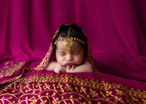 Sleeping newborn baby peeking from ornate gold-embroidered magenta fabric with traditional Indian headpiece.