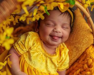 Smiling newborn baby girl in yellow dress with bow headband lying in basket with yellow flowers.