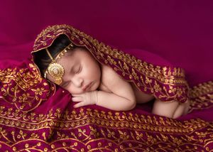 Peaceful newborn baby wrapped in ornate gold and magenta Indian bridal fabric with traditional jewelry.