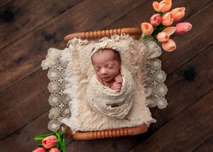 Sleeping newborn baby wrapped in cream lace swaddled in a tiny wooden bed surrounded by pink tulips.