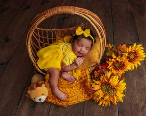 Sleeping newborn baby in yellow dress with bow in wicker basket with sunflowers and teddy bear.
