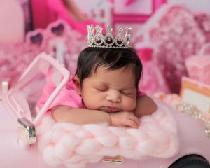 Newborn baby girl wearing a sparkly crown sleeping in a pink toy car with fluffy bedding.