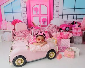 Sleeping newborn baby in pink outfit posed in toy convertible car in Barbie-themed pink studio.