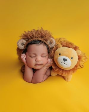 Cute newborn baby in lion costume hat sleeping beside a stuffed lion toy on yellow backdrop.