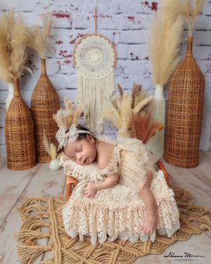 Sleeping newborn baby in cream lace dress on ornate chair with boho dreamcatcher decor backdrop.