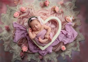 Newborn baby curled in heart-shaped frame on purple and lace blanket with pink roses.