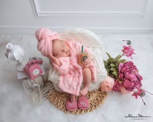 Newborn baby sleeping in pink fluffy robe and hat in a wicker basket with flowers and props.
