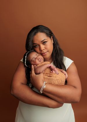 Mother in white dress tenderly holding sleeping newborn against brown background.