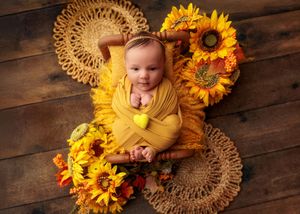 Smiling baby wrapped in yellow swaddle holding a felt heart surrounded by sunflowers on wood floor.
