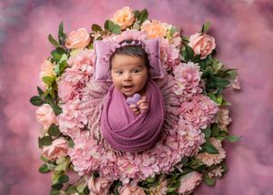 Smiling newborn wrapped in purple knit swaddle and bonnet nestled in a ring of pink hydrangeas.