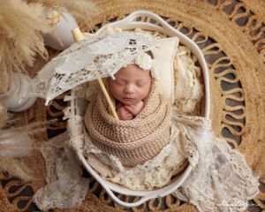 Newborn baby swaddled in knit wrap holding a lace umbrella in a boho-styled wicker basket.