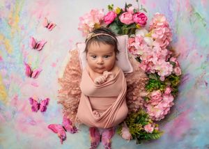 Newborn baby in pink wrap surrounded by pink flowers and butterfly decorations on colorful background.