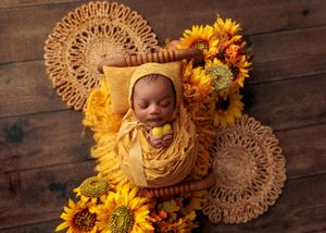 Newborn baby in yellow bonnet swaddled in wooden bowl surrounded by vibrant sunflowers.