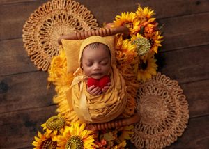 Sleeping newborn in yellow bonnet and wrap lying in wooden bed with sunflower arrangement.