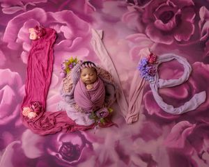 Newborn baby wrapped in purple swaddle posed inside LOVE letters on vibrant floral pink background.