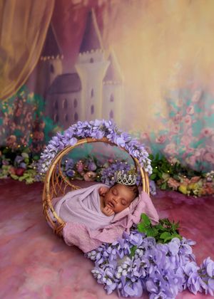 Sleeping newborn wrapped in lavender in wicker basket with purple flowers and castle backdrop.